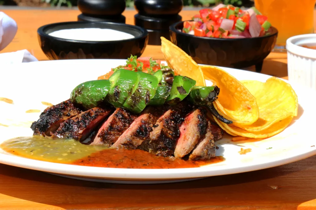 Sliced grilled carne asada topped with grilled peppers, served with tortilla chips and red and green salsa on a white plate.