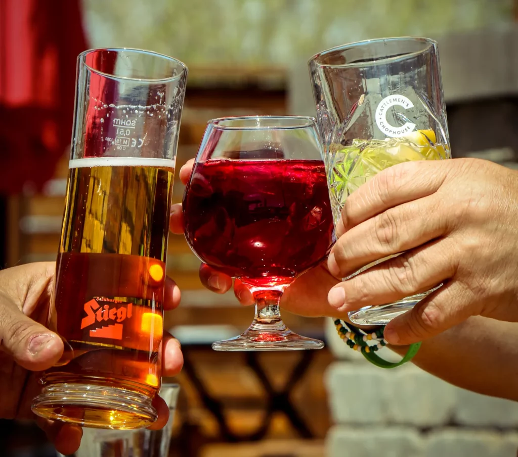 Three hands clink glasses together on a sunny patio—one with a golden beer, one with a red cocktail, and one with a clear drink garnished with lemon.